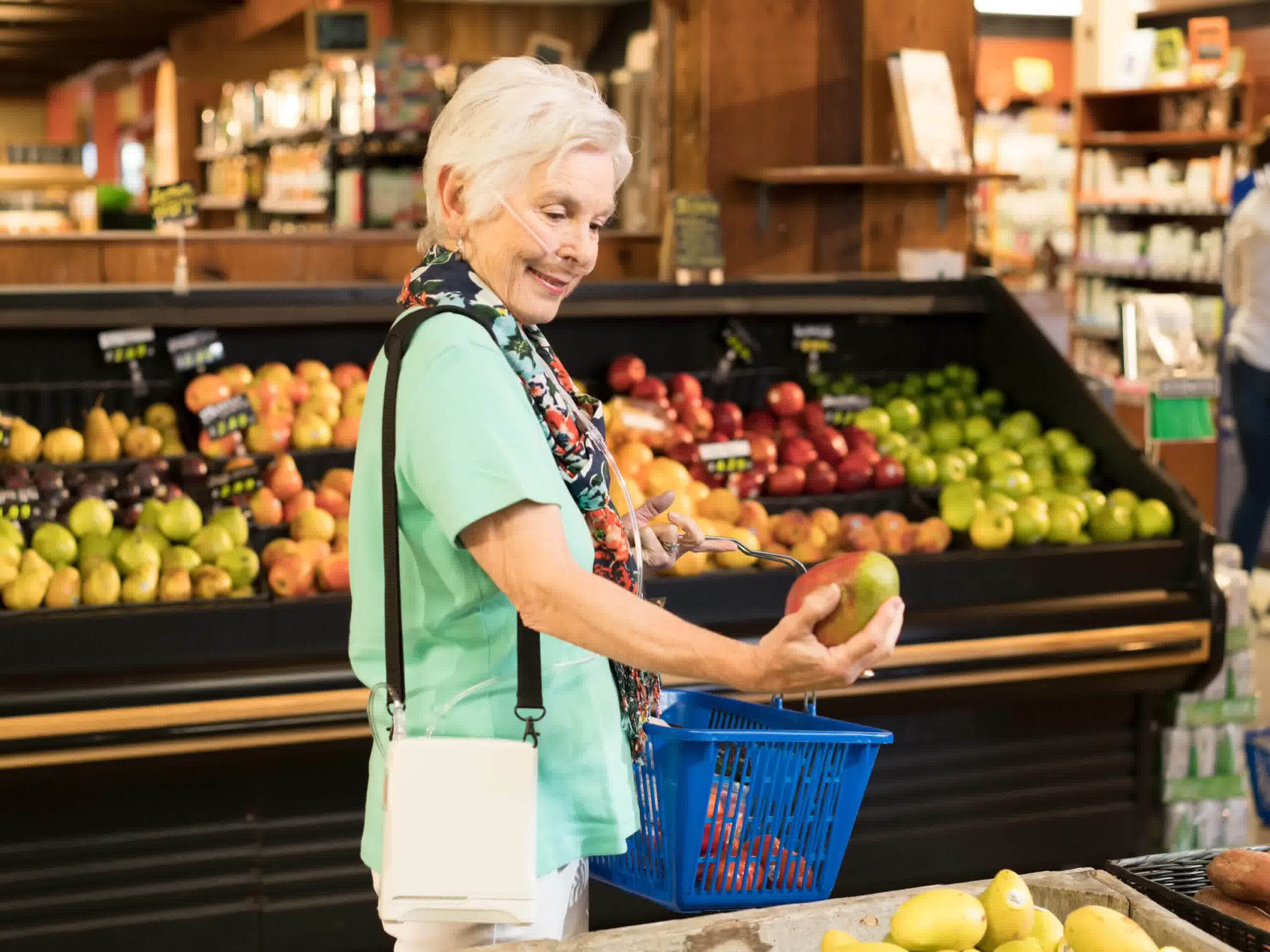 An older woman with a shopping basket examines a mango in the produce section of Aspen Concentrators grocery store, surrounded by colorful fruits like apples and pears. For inquiries, please Contact Us.