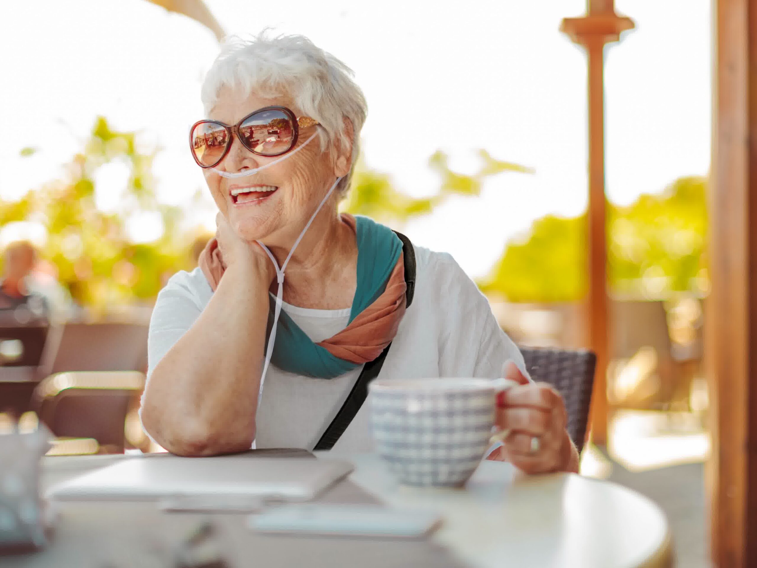 Smiling older woman with short white hair and sunglasses sits outdoors at a table, holding a checkered mug. She wears a colorful scarf and white shirt, with an Aspen Concentrators oxygen tube, enjoying a sunny day. Contact Us to learn more.