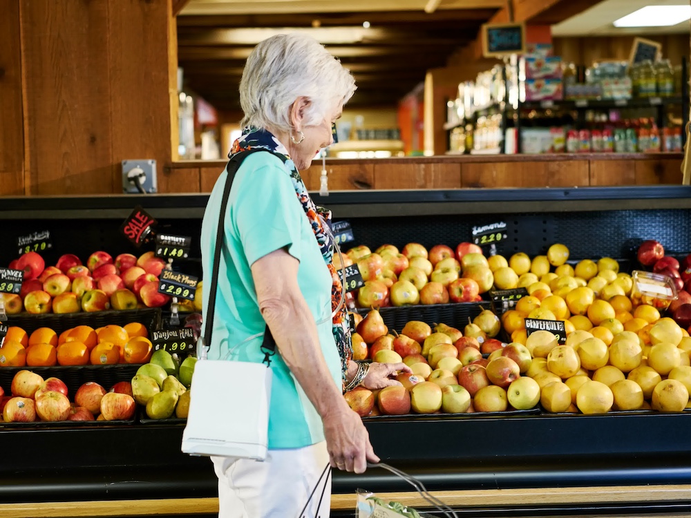 An elderly woman with short gray hair shops for apples in a grocery store, smiling as she holds a basket and examines fruit—perhaps after visiting an Oxygen Concentrator Repair Colorado Springs service nearby.